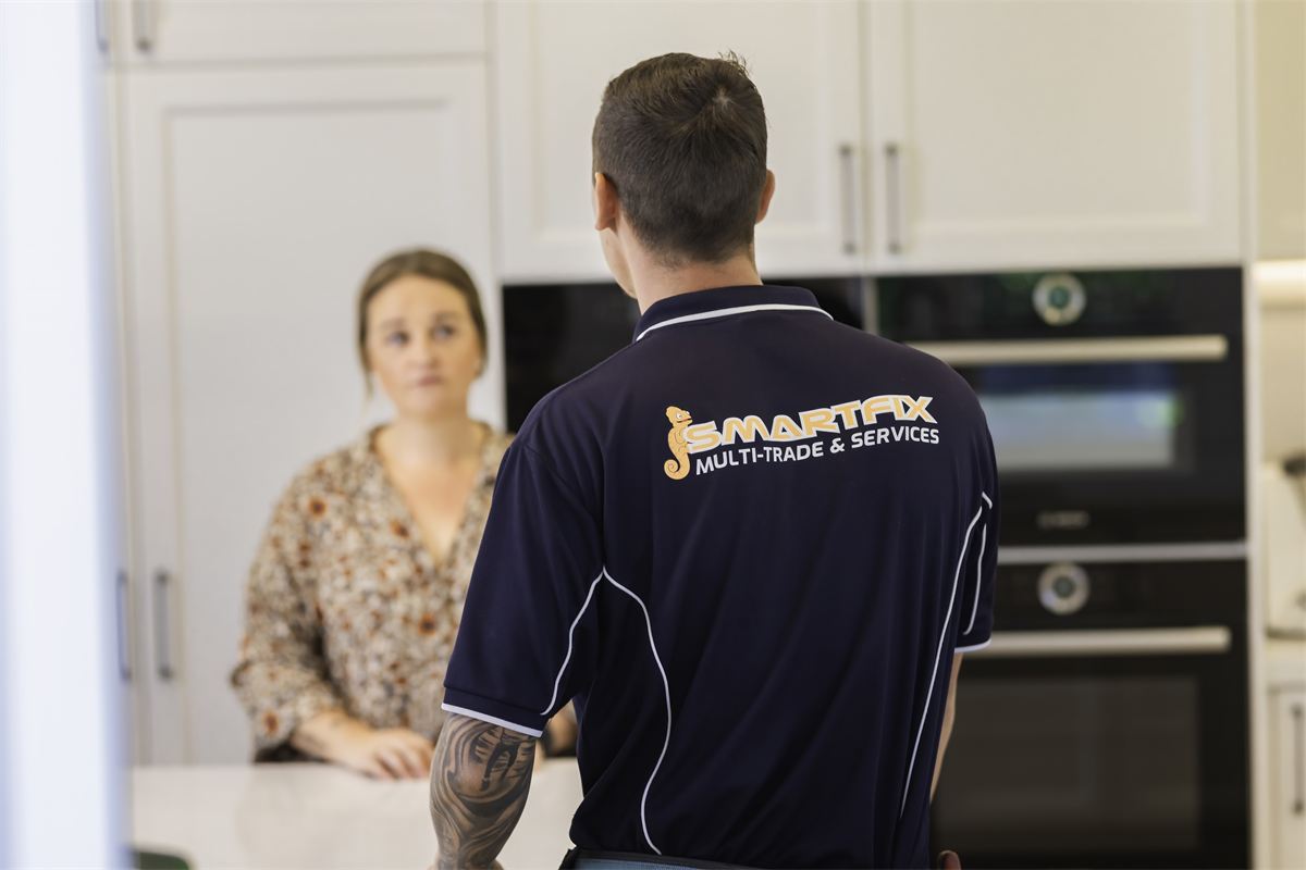 Smartfix technician in a uniform with Colin on the back, talking with a homeowner in her kitchen